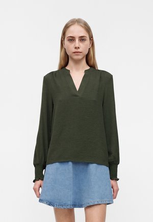 Jeune femme debout, tournant vers l'avant, portant un chemisier vert foncé à manches longues avec un col en V et une jupe en jean bleu clair.