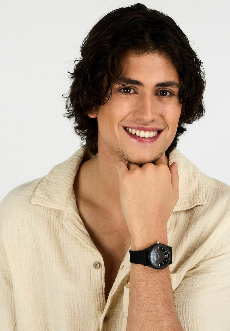 Orologio digitale nero con quadrante quadrato e accenti rossi, indossato su un polso. Il cinturino è in silicone, in abbinamento a una camicia beige texturizzata.