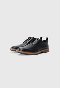 Zapatos de cuero negro con punta redondeada, diseño de cordones y una suela texturada marrón que destacan una estética moderna y minimalista.