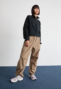 Pulover negru din fleece cu guler cu fermoar pe jumătate, purtat cu pantaloni cargo bej largi și adidași violeți deschiși cu accente gri.