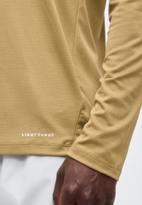 Κοντινό σε βραχίονα και χέρι ατόμου που φοράει μια υφασμάτινη ανοιχτοκάστανη μπλούζα με μακριά μανίκια, με την ετικέτα "LIGHTRANGE" και είναι ορατό το λευκό παντελόνι.