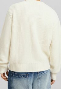 Man som bär en krämfärgad ribbad stickad tröja och ljusblå denimjeans, sedd bakifrån mot en enfärgad bakgrund.