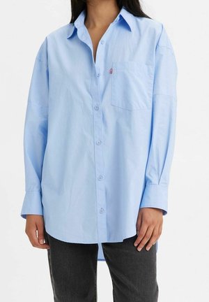 Femme portant une chemise bleu clair oversize avec poche poitrine et pantalon noir, debout devant un fond uni.
