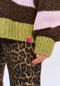 Gruby sweter we wzór pasków w odcieniach brązu, różu i zieleni, z ściągaczami na rękawach. Nosi się go z spodniami w panterkę, prezentując różowy detal metki.