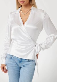 Balta satino bluzė su giliu V formos kaklu, ilgomis rankovėmis ir šoniniu raišteliu. Suderinta su šviesiai mėlynomis džinsomis.
