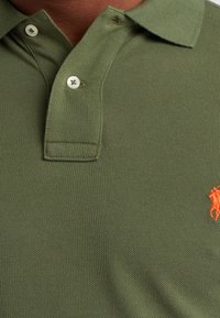 Polo vert olive en tissu tricoté texturé, avec une patte de boutonnage à trois boutons et un petit logo de cheval orange sur la poitrine gauche.