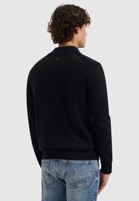 Mann mit lockigem, dunklem Haar, der einen schwarzen Pullover mit langen Ärmeln und blaue Jeans trägt, von hinten vor einem einfarbigen grauen Hintergrund gezeigt.