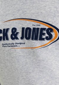 Maglietta grigia in tessuto con grande testo blu che recita "Jack & Jones" e una linea accentuata arancione. Include il testo "Est. 1990" e "Design Estetico".