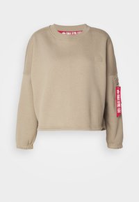 Beige cropped sweatshirt with long sleeves and elastic cuffs. Features a small logo patch and a red tag reading "REMOVE BEFORE FLIGHT."