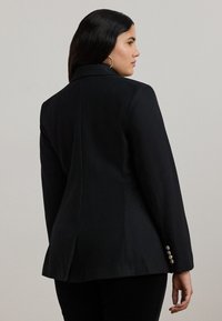 Fekete, testhez simuló blézer hajtókával, strukturált kialakítással, szatén textúrával és négygombos mandzsetta részlettel. Sötét nadrág fölött viselve.
