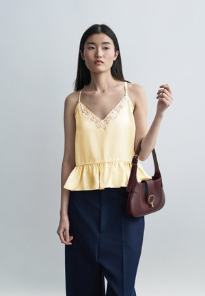 Jeune femme aux longs cheveux bruns portant un haut jaune sans manches orné de dentelle et un pantalon bleu marine, tenant un petit sac à main marron.