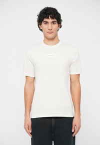 Man with curly hair wearing a white Armani Exchange t-shirt and black pants, standing against a plain light background.