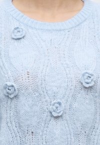 Pull en tricot bleu clair avec des motifs floraux en relief et de petites fleurs tricotées en 3D sur le devant, porté par une personne.