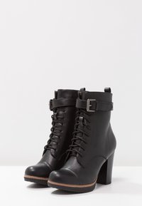 Botas de piel negra hasta el tobillo con tacón alto, con cordones en la parte delantera, correa decorativa con hebilla, punta redonda y superficie texturizada.