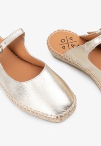 Zapatos espadrilles de color plateado metálico con punta redondeada, correa ajustable y suela de yute trenzado. Textura suave con detalle de costura en contraste.