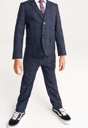 Costume à carreaux bleu marine avec deux boutons, revers crantés et poches, associé à des baskets noires et blanches. Coupe ajustée et tissu texturé.