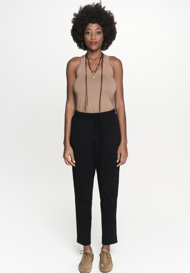 Brązowy top na ramiączkach, czarne spodnie i beżowe buty. Modelka z kręconymi włosami nosząca długi naszyjnik. Prosty design i neutralne kolory.