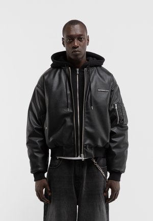 Mann trägt eine schwarze Kapuzenjacke unter einer schwarzen Lederjacke mit Reißverschluss und einer Kettenverzierung auf schwarzen Hosen.