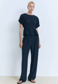 La modella si trova di fronte a uno sfondo chiaro, indossando un top blu scuro e un pantalone ampio abbinato, con sandali e un'espressione neutra.