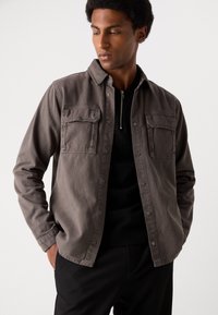 Camisa gris de botones, hecha de una tela texturizada, que cuenta con dos bolsillos en el pecho y un cuello. Se lleva sobre una parte superior negra, con un corte relajado.