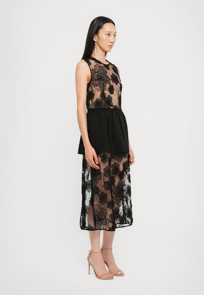 Vestido negro de encaje con bordado floral, diseño sin mangas, cintura tipo peplum, tela transparente, dobladillo a la altura de la rodilla, combinado con tacones nude con tiras.