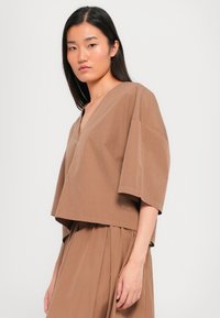 Brun cropped topp med korte ermer, V-hals, glatt tekstur og subtilt merketag på siden. Brukt med et matchende skjørt.