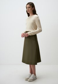 Crème lange mouwen trui met geribbelde manchetten, gecombineerd met een olijfgroene A-lijn rok. Witte en beige sportschoenen maken de look compleet.
