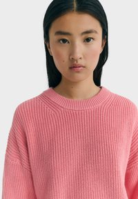 Jeune femme asiatique avec des cheveux noirs raides, portant un pull en tricot rose texturé, faisant face à l'avant avec une expression neutre sur un fond uni.