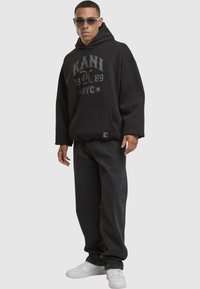 Sudadera negra con la impresión "KANI 1989 NYC" en gris, combinada con jeans oscuros y zapatillas blancas. Presenta un bolsillo frontal y detalle del logo.