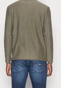 Chemise à manches longues vert olive avec une surface texturée et une coupe droite, associée à un jean en denim bleu avec des poches arrière et des détails de couture.