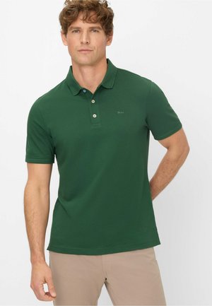 Man with curly hair wearing a dark green short-sleeve polo shirt and beige pants, standing with one hand behind his back.