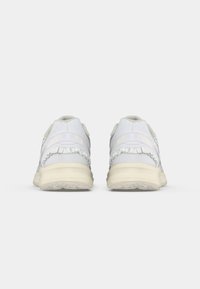 Weiße Sportschuhe mit einer strukturierten Oberseite, gerafften Akzenten an der Ferse und einer cremefarbenen Sohle. Die Rückansicht zeigt eine gepolsterte Kragenpartie und Nähdetails.