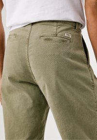 Vue de dos d'une personne portant un pantalon vert olive avec des poches passepoilées à l'arrière et une chemise blanche rentrée.