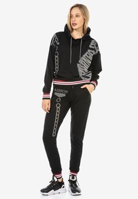 Schwarzes Hoodie- und Jogginganzug-Set mit Kapuze, gestreiften Akzenten und dekorativen silbernen Kettenmotiven. Weicher Stoff, lockere Passform und gerippte Bündchen.