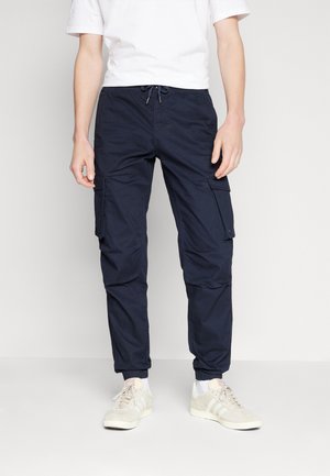 Homme portant un pantalon cargo bleu marine avec des poignets élastiques, des baskets blanches et une chemise blanche à manches courtes, debout devant un fond uni.