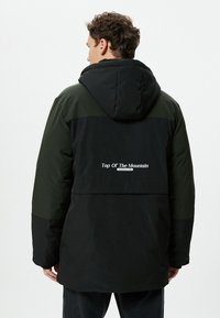 Schwarz-grüne Kapuzenjacke mit lockerer Passform. Sie verfügt über ein Logo auf dem Rücken, kontrastierende Einsätze und eine strukturierte Außenseite.