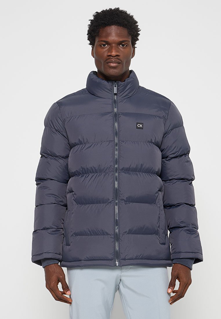 Homme portant une veste matelassée gris foncé avec fermeture éclair à l'avant et logo CK sur la poitrine, se tenant devant un fond uni.