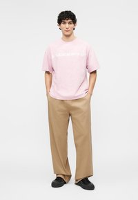 Jeune homme aux cheveux bouclés portant un t-shirt rose "ENDORPHIN", un pantalon beige à jambes larges et des baskets noires, debout les mains dans les poches.