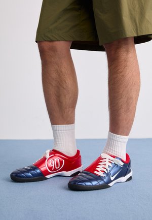 Las zapatillas deportivas rojas y marinas presentan una textura acolchada, cordones blancos y un gran logotipo '90'. Se llevan con calcetines blancos y pantalones cortos verdes.