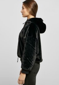 Čierny skrátený hoodie vyrobený z jemného, plyšového materiálu. Obsahuje kapucňu so šnúrkou, elastické manžety a voľný strih.
