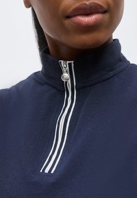 Κοντινό πλάνο ατόμου που φοράει μπλε σκούρο φούτερ με φερμουάρ με λευκές ρίγες και στρογγυλό ασημένιο τράβηγμα φερμουάρ.