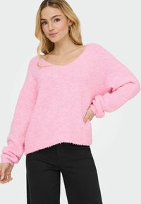 Rožna, puhasta pletena jopica z širšim izrezom in dolgimi rokavi, z odrezanim robom in teksturirano tkanino. V kombinaciji z črnimi hlačami.