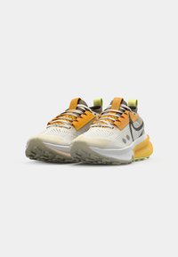Hardloopschoenen met een beige mesh bovenkant, oranje en grijze accenten, en een gevoerde kraag. Voorzien van ZoomX demping en een gestructureerde rubberen zool.