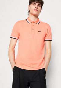 Pfirsichfarbendes Poloshirt mit einem kontrastierenden schwarz-weißen gestreiften Kragen, einer Drei-Knopf-Leiste und einem gestickten Logo auf der Brust.