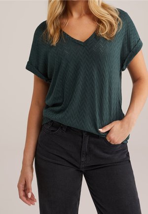 Vrouw draagt donkergroene, gestructureerde korte mouw V-hals top en zwarte high-waist jeans met één hand in de zak, tegen een effen achtergrond.