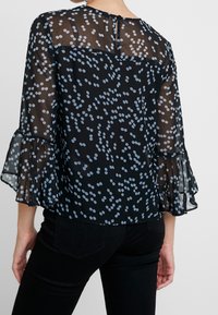 Schwarze, durchsichtige Bluse mit einem gepunkteten Muster in Hellblau. Verfügt über weit ausgestellte Ärmel und einen geknöpften Schlitz im Rücken. Leichte Textur.