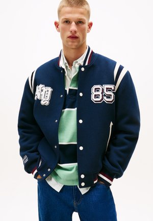 Giacca varsity navy con strisce bianche e bordeaux, toppe ricamate e un'esterno in lana. Indossata sopra una camicia a righe verdi e blu.