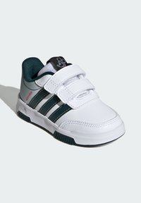 Sneaker bianche con accenti verdi, dotate di cinturini in velcro, una tomaia in pelle testurizzata e una linguetta nera con un design di un personaggio.