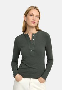 Ciemnozielona, prążkowana bluzka z długim rękawem, z kołnierzykiem henley, plisą z guzikami z pięcioma guzikami oraz dopasowanym fasonem.
