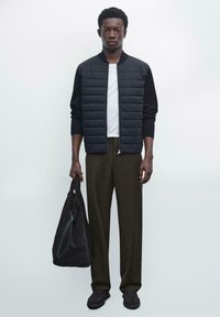 Giacca bomber nera imbottita sopra una maglietta bianca, pantaloni verde oliva e una borsa a tracolla in suede scuro. Presenta sneakers scure e uno sfondo neutro.